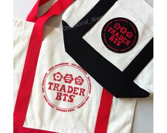 Puede incluir: Un bolso de tela blanco con correas y base rojas, con un logotipo circular rojo que dice TRADER BTS rodeado de nombres. Un bolso de tela más pequeño con ribete negro y un parche circular negro con el texto en rojo TRADER BTS.