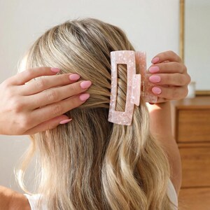 Puede incluir: Una pinza para el pelo rosa claro con forma rectangular y diseño veteado. El accesorio para el cabello se coloca en cabello rubio. La pinza es un accesorio elegante para varios peinados.