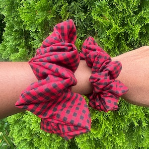 May include: Two fabric hair scrunchies featuring a red and black buffalo plaid checkered pattern. The scrunchies are gathered with elastic, creating a ruffled, voluminous appearance. The fabric has a visible woven texture throughout the design.