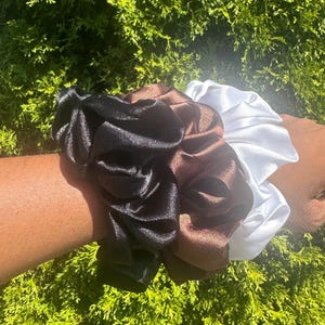 May include: Three satin scrunchies in black, brown, and white. The scrunchies are gathered and have a shiny, smooth texture. The scrunchies are displayed against a backdrop of green foliage.