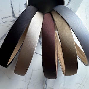 May include: Six different colored satin headbands. The headbands are arranged in a circle, with the black headband at the top and the gray headband at the bottom. The headbands are all the same size and width.