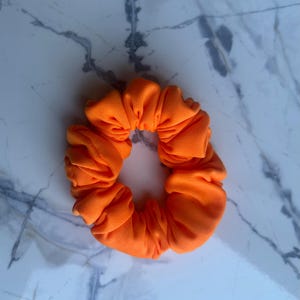 Könnte beinhalten: Hellorangefarbenes Haargummi aus Stoff. Das Haargummi ist kreisförmig und gerafft, mit einer leuchtenden, einfarbigen Farbe. Der Stoff scheint ein weiches Material zu sein. Ein Haaraccessoire.