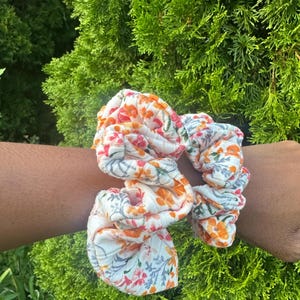 May include: A white ribbed fabric hair scrunchie featuring a repeating floral pattern in shades of orange, pink, and muted green. The accessory has a gathered, ruffled texture and is shown as a circular band with a soft, flexible construction.