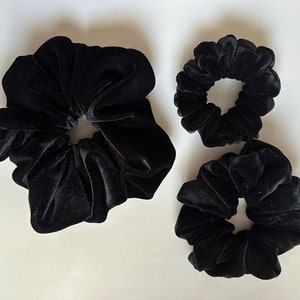 May include: Three black velvet scrunchies of different sizes. The largest scrunchie is in the center of the image, and the two smaller scrunchies are on either side of it.