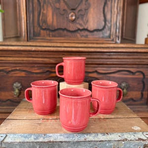 Könnte beinhalten: Vier rote Keramikbecher mit Henkeln stehen auf einer Holzoberfläche. Ein Becher steht auf einem kleinen, quadratischen, weißen Block. Die Becher sind einfarbig, leuchtend rot und scheinen von ähnlicher Größe und Form zu sein.