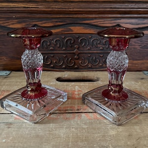 Könnte beinhalten: Zwei Vintage-Kerzenhalter aus Glas. Jeder hat eine rubinrote Oberseite und einen Sockel mit einem klaren, rautenförmigen Stiel. Die Ränder sind mit Gold verziert. Die Kerzenhalter stehen auf einer quadratischen, klaren Glasbasis.