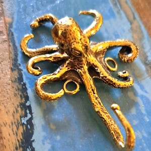 May include: A gold-colored octopus hook with a textured surface. The hook is designed to resemble an octopus with eight tentacles and a curled tail.