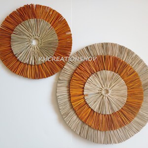 May include: Two round wall decor pieces made of woven natural materials. The larger piece is beige with a brown outer ring. The smaller piece is brown with a beige outer ring.
