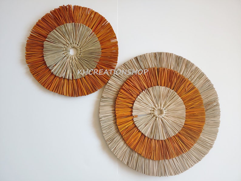 May include: Two round wall decor pieces made of woven natural materials. The larger piece is beige with a brown outer ring. The smaller piece is brown with a beige outer ring.