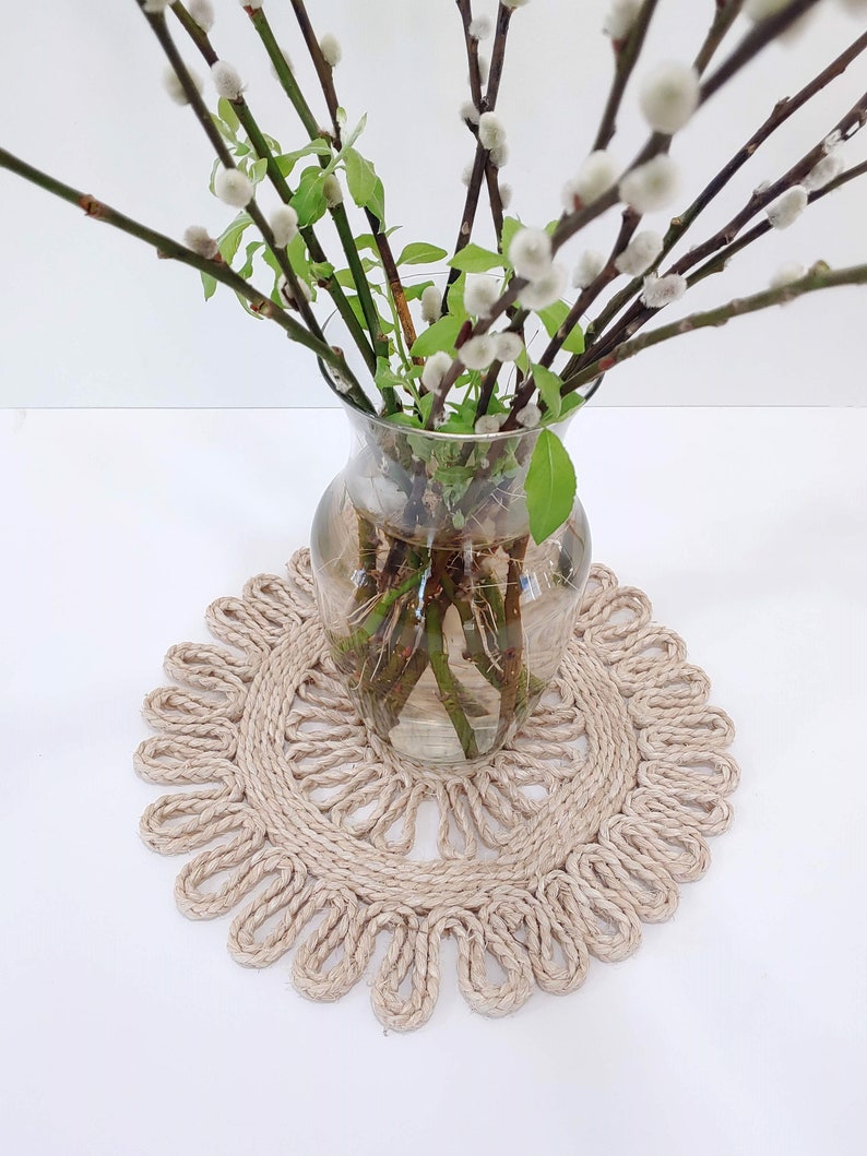 May include: A glass vase with pussy willow branches in it, sitting on a round, beige, woven coaster.