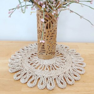May include: A natural woven jute placemat with a scalloped edge. The placemat is beige and has a delicate, intricate design. A carved wooden vase with a floral pattern is sitting on top of the placemat.