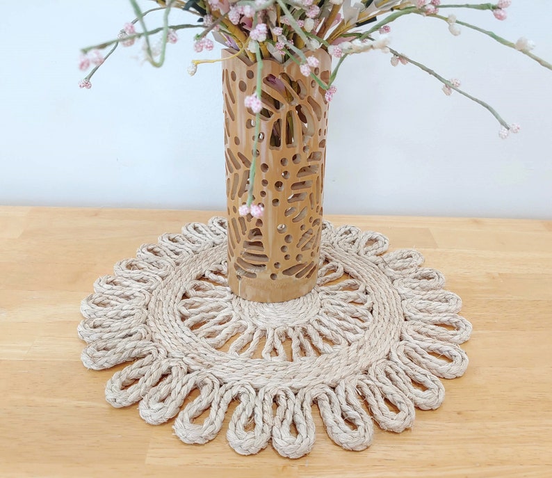 May include: A natural woven jute placemat with a scalloped edge. The placemat is beige and has a delicate, intricate design. A carved wooden vase with a floral pattern is sitting on top of the placemat.