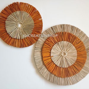 May include: Two round wall decor pieces made of woven natural materials. The larger piece has three concentric circles, the outer two are orange and the inner one is beige. The smaller piece has two concentric circles, the outer one is orange and the inner one is beige.