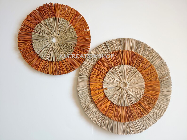 May include: Two round wall decor pieces made of woven natural materials. The larger piece has three concentric circles, the outer two are orange and the inner one is beige. The smaller piece has two concentric circles, the outer one is orange and the inner one is beige.
