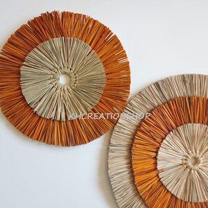 May include: Two round wall decor pieces made of woven natural materials. The larger piece is in the background and the smaller piece is in the foreground. Both pieces have a concentric circle design with a light brown center and an outer ring of orange.