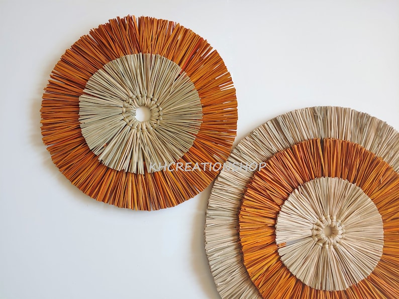 May include: Two round wall decor pieces made of woven natural materials. The larger piece is in the background and the smaller piece is in the foreground. Both pieces have a concentric circle design with a light brown center and an outer ring of orange.