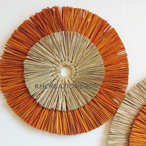 May include: Two round wall decor pieces made of woven natural materials. The larger piece has a center circle of natural straw with an outer ring of orange straw. The smaller piece has a similar design but is smaller.