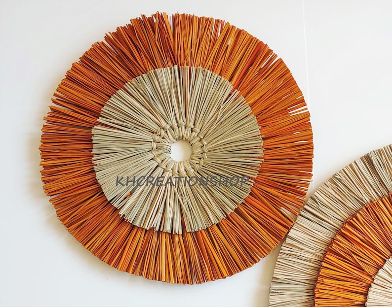 May include: Two round wall decor pieces made of woven natural materials. The larger piece has a center circle of natural straw with an outer ring of orange straw. The smaller piece has a similar design but is smaller.