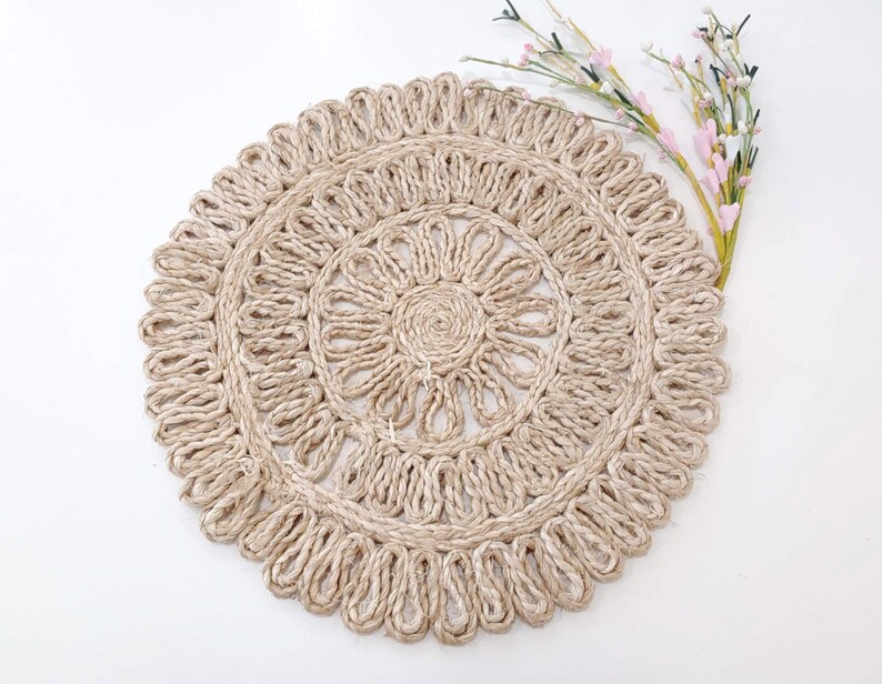 May include: A round, beige, woven placemat with a decorative, floral design. The placemat is made of natural fibers and has a rustic, bohemian style.