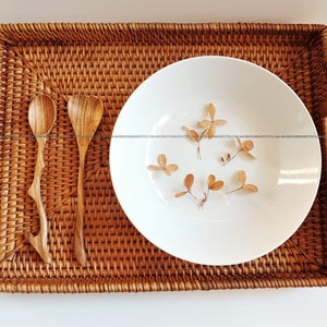 May include: A white ceramic bowl with dried flowers sits on a woven rattan tray with two wooden spoons.