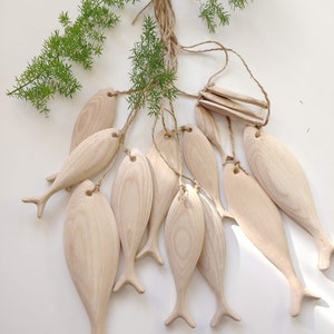 K&ouml;nnte beinhalten: Eine Sammlung von hellen Holzfisch-Anh&auml;ngern, die jeweils mit einer glatten Oberfl&auml;che und sichtbarer Holzmaserung geschnitzt sind. Die Fische sind an einem zentralen Punkt mit d&uuml;nnen Juteschn&uuml;ren befestigt und bilden ein B&uuml;ndel h&auml;ngender Formen.