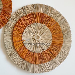 May include: A large, round wall decor made of natural materials. It features three concentric circles, with the outer circle being a light beige, the middle circle being a bright orange, and the inner circle being a light beige. The circles are made of woven straw-like material.