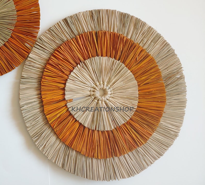 May include: A large, round wall decor made of natural materials. It features three concentric circles, with the outer circle being a light beige, the middle circle being a bright orange, and the inner circle being a light beige. The circles are made of woven straw-like material.