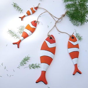 K&ouml;nnte beinhalten: Ein Set aus vier flachen, h&ouml;lzernen Fischornamenten, die mit orangefarbenen und wei&szlig;en Streifen bemalt sind. Jeder Fisch hat ein kleines Loch an der Oberseite, durch das eine nat&uuml;rliche Jutekordel zum Aufh&auml;ngen gezogen ist.