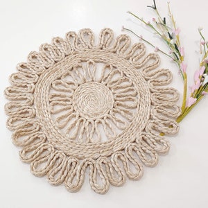 May include: A round, beige, crocheted placemat with a delicate, floral design. The placemat is made of natural fibers and has a rustic, bohemian aesthetic.