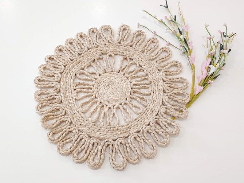 May include: A round, beige, crocheted placemat with a delicate, floral design. The placemat is made of natural fibers and has a rustic, bohemian aesthetic.