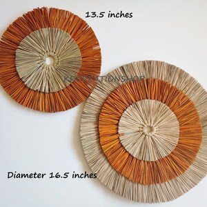 May include: Two round wall decor pieces made of woven natural materials. The larger piece is 16.5 inches in diameter and the smaller piece is 13.5 inches in diameter. Both pieces have a natural beige color with orange accents.