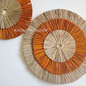 May include: Two round wall decor pieces made of woven natural materials. The larger piece has three concentric circles, the inner two are orange and the outer one is natural. The smaller piece has two concentric circles, the inner one is natural and the outer one is orange.