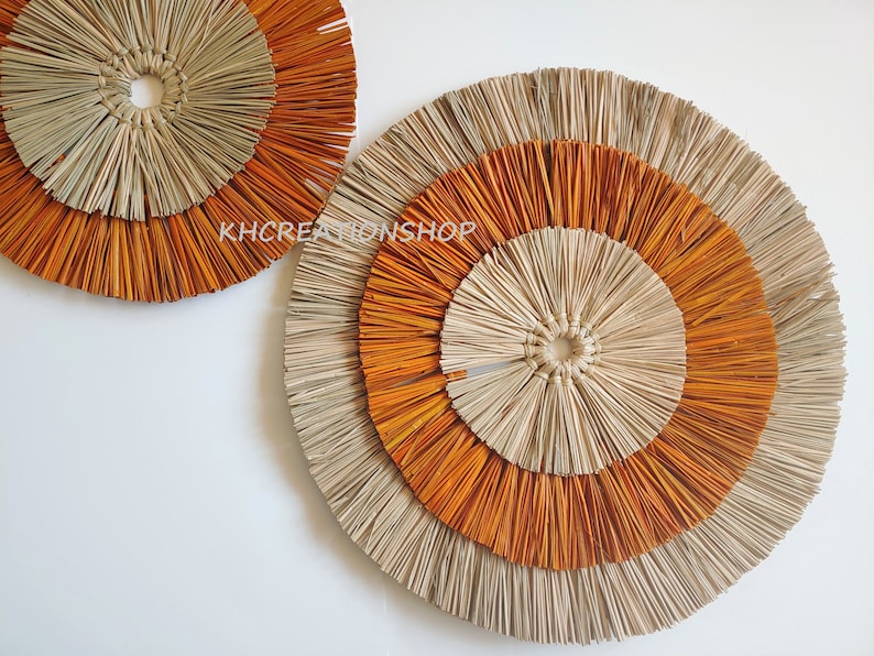 May include: Two round wall decor pieces made of woven natural materials. The larger piece has three concentric circles, the inner two are orange and the outer one is natural. The smaller piece has two concentric circles, the inner one is natural and the outer one is orange.