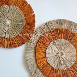 May include: Two round wall decor pieces made of woven natural materials. The larger piece has three concentric circles, the outer two are orange and the inner one is beige. The smaller piece has two concentric circles, the outer one is orange and the inner one is beige.