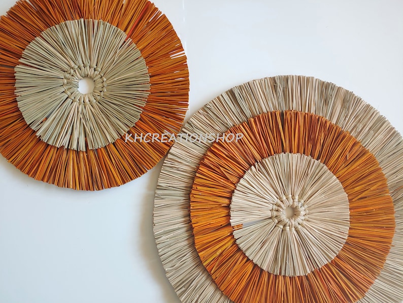 May include: Two round wall decor pieces made of woven natural materials. The larger piece has three concentric circles, the outer two are orange and the inner one is beige. The smaller piece has two concentric circles, the outer one is orange and the inner one is beige.