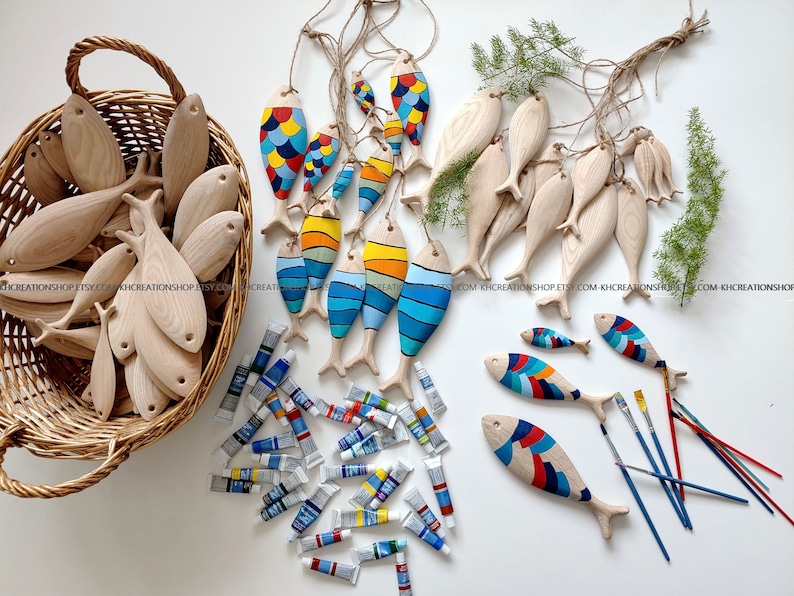 K&ouml;nnte beinhalten: Eine Sammlung von Fischornamenten aus Holz, teils naturbelassen, teils handbemalt mit bunten Mustern in Blau, Gelb, Rot und Orange. Das Set enth&auml;lt einen Weidenkorb, zahlreiche Farbtuben und mehrere d&uuml;nne Pinsel f&uuml;r kreative Arbeiten.