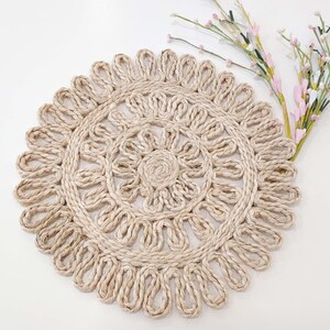 May include: A round, beige, woven placemat with a decorative, intricate design. The placemat is made of natural fibers and has a delicate, lacy appearance.