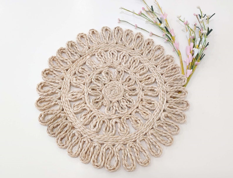 May include: A round, beige, woven placemat with a decorative, intricate design. The placemat is made of natural fibers and has a delicate, lacy appearance.