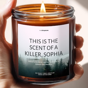 May include: A lit soy wax candle in a glass jar. The label reads "THIS IS THE SCENT OF A KILLER, SOPHIA." The candle has a forest scene and the text "Infused with charm, mystery, and a hint of Edward-level obsession."