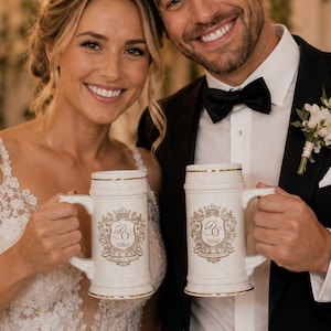 May include: Two white ceramic beer steins featuring a gold-toned crest design with the initials DG in the center. Each stein has a sturdy handle, a textured base, and a gold rim. The mugs are held by two individuals dressed in formal wedding attire.