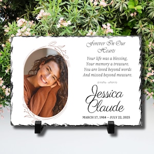 May include: A rectangular slate memorial plaque with a rough, chiseled edge. It features a portrait of a smiling person inside an oval frame, with the text Forever In Our Hearts, a poem, the name Jessica Claude, and the dates March 17, 1984 – July 22, 2025.