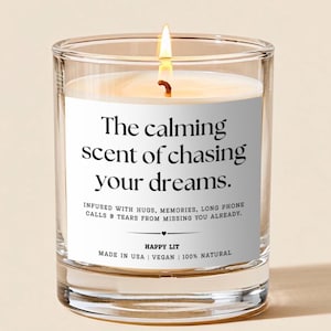 May include: A clear glass candle with a lit wick and a white label featuring the black text: The calming scent of chasing your dreams. Infused with hugs, memories, long phone calls & tears from missing you already. Happy Lit. Made in USA | Vegan | 100% Natural.