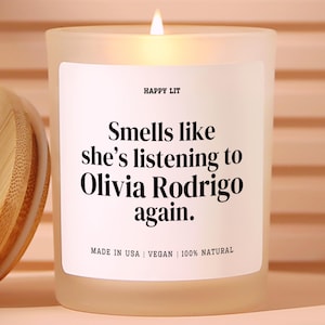 May include: A frosted glass candle with a lit wick and a white label featuring the text HAPPY LIT, Smells like she's listening to Olivia Rodrigo again., MADE IN USA | VEGAN | 100% NATURAL. A bamboo lid is positioned to the left of the candle.