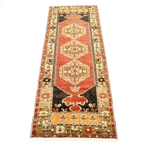 May include: A long rectangular runner rug featuring a central terracotta red field with three cream-colored geometric medallions. The border displays intricate floral and geometric patterns in shades of black, tan, orange, and cream on a dark background.