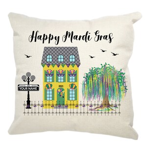 May include: A decorative pillow with a yellow house with green shutters and a purple, green, and gold Mardi Gras mask on the door. The house has a gray roof and is surrounded by a black fence. There is a purple, green, and gold tree with purple flowers hanging down. The pillow has the text "Happy Mardi Gras" and "YOUR NAME" on it.