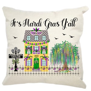 May include: A decorative pillow with a yellow house with purple and green accents. The house has a sign that says "Taylor" and the year "2023". The text "It's Mardi Gras Y'all" is written in black cursive. The pillow has a white background.