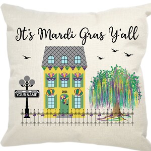 May include: A yellow and green house with a gray roof and purple and green Mardi Gras beads hanging from the windows. The house has a green door with a jester's face on it. The year 2021 is written on the door. A sign in front of the house says "YOUR NAME". A purple and green weeping willow tree is in front of the house. The text "It's Mardi Gras Y'all" is at the top of the image.