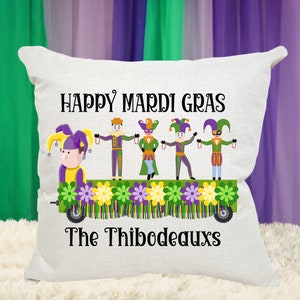 May include: White throw pillow with the text "Happy Mardi Gras" and a cartoon image of a Mardi Gras parade float with five people in jester costumes. The text "The Thibodeauxs" is below the float.