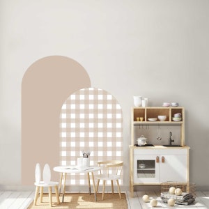 May include: A wooden play kitchen with white and natural wood elements, alongside a small table and chairs. The wall features a beige arch design. The scene is completed with a rug and decorative balls.