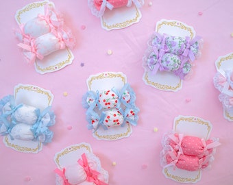 May include: A collection of fabric hair accessories shaped like wrapped candies, each mounted on a white card with gold trim. The accessories feature various patterns including cherries, polka dots, and floral prints in pink, blue, and purple.